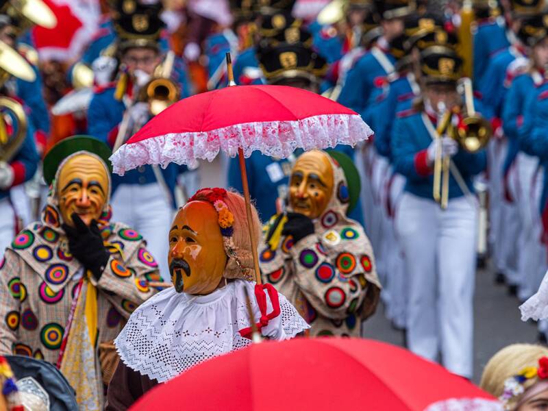 Abgebildet wird ein Narrensprung in Oberndorf am Neckar. Im Zentrum des Bildes ist der Hansel mit rotem Schirm zu erkennen. Der Hansel ist einer der Hauptfiguren der Oberndorfer Fasnet. Hinter dem Hansel sind zwei Schantles zu erkennen. Der Schantle ist eine weitere Hauptfigur der Oberndorfer Fasnet. Hinter den Narren ist die Musikkapelle zu sehen.