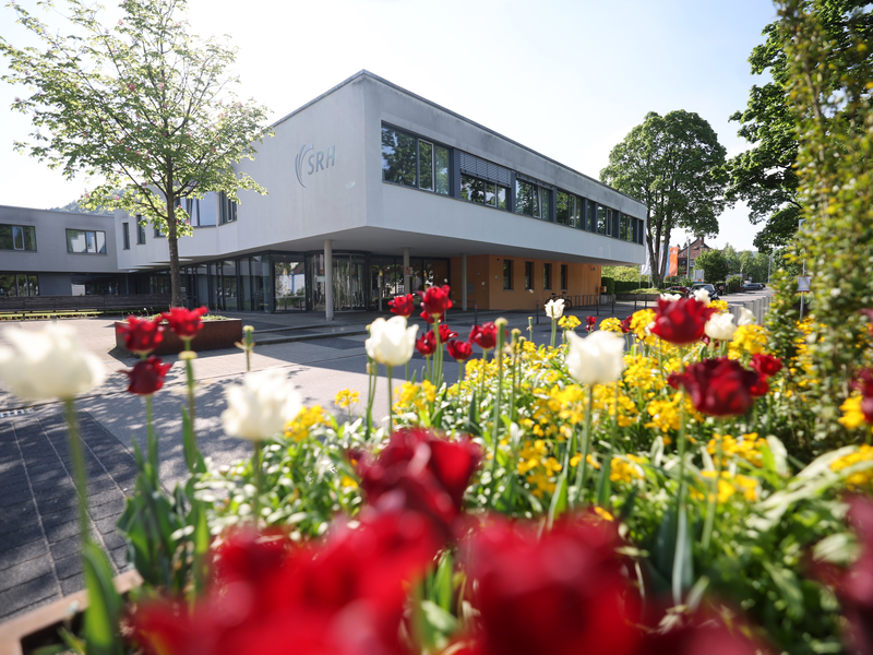 Abgebildet ist das SRH Krankenhaus in Oberndorf am Neckar. Im Vordergrund sind bunte Blumen zu erkennen.