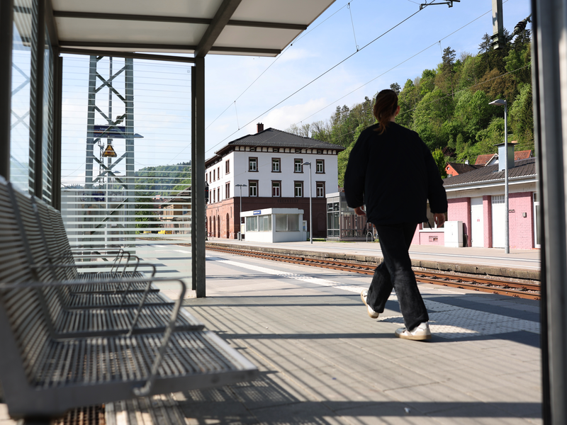 Abgebildet wird der Bahnhof in Oberndorf am Neckar. Eine junge Frau läuft neben den Schienen den Banhof entlang.