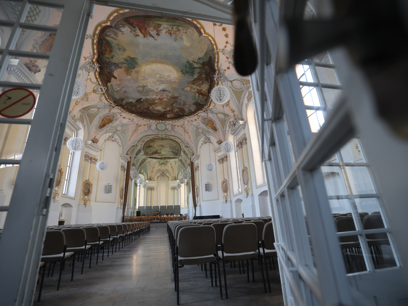Ein Blick vom Eingang aus in die Klosterkirche. Stuhreihen links und rechts.  Umfangreiche Deckenmalereien. Blick auf die Tribüne.