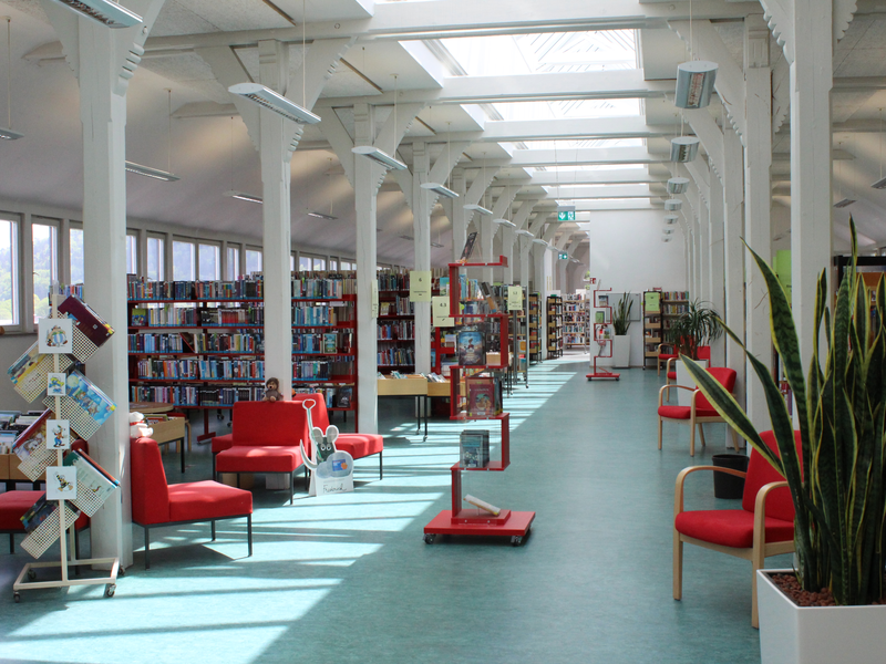 Ein lichtdurchfluteter Gang mit weißen Säulen und vielen Bücherregalen. Dazwischen stehen rote Sitzgelegenheiten.