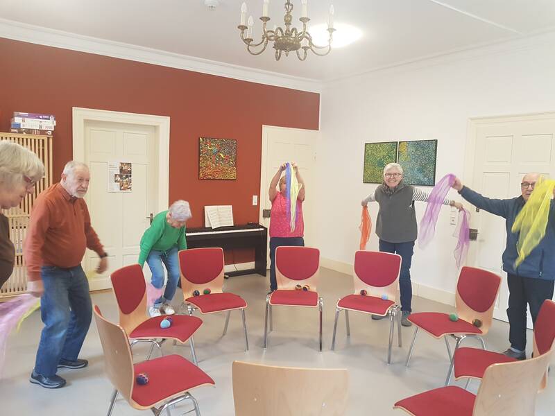 Ein Struhlkreis ist in der Mitte. Drumherum stehen Rentner und Rentnerinnen und machen mit bunten Tüchern Gymnastik. 