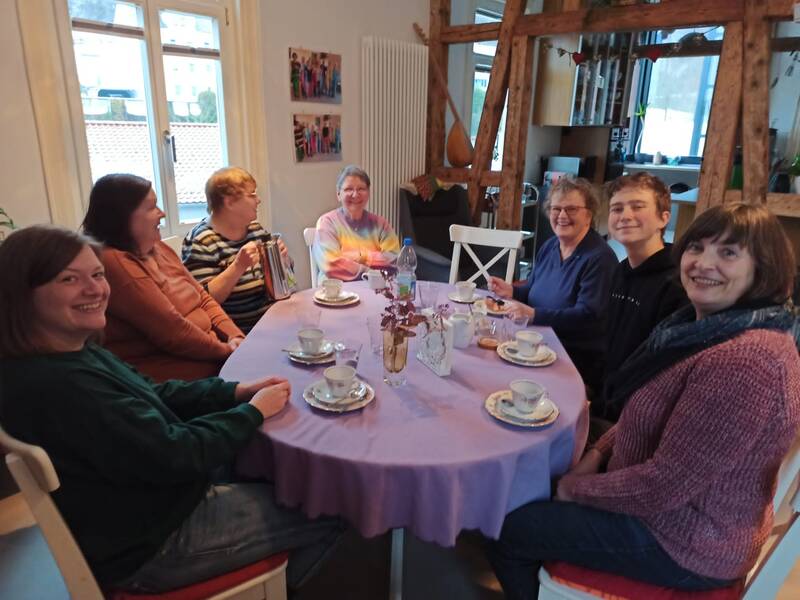6 Damen und ein junger Herr sitzen um einen Kaffeetisch. Auf dem Tisch steht älteres Kaffeeporzellan. Alle lächeln freundlich in die Kamera.