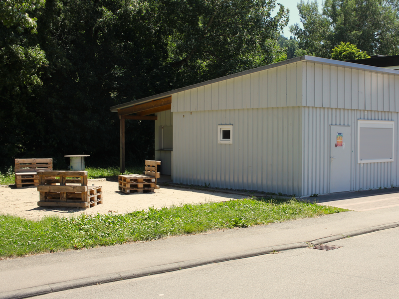 Ein kleines grauses einstöckiges Gebäude welches fast quadratisch ist. Auf dem kleinen Vorplatz stehen drei Sitzmöglichkeiten aus Holzpaletten. 