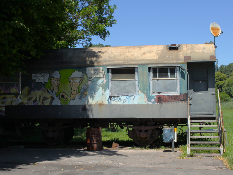 Ein mit Graffiti bemalter älterer Bauwagen mit Schornstein und Treppenaufgang. Hinter dem Baugwagen beginnt eine Wiese. Der Bauwagen steht auf einem geteerten Platz. 