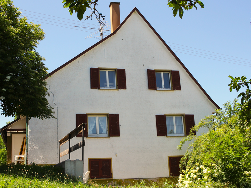 Rückansicht aus dem Garten heraus auf ein Haus mit 4 offenen und zwei geschlossenen Fenster. Im Garten befinden sich Büsche und ein Baum
