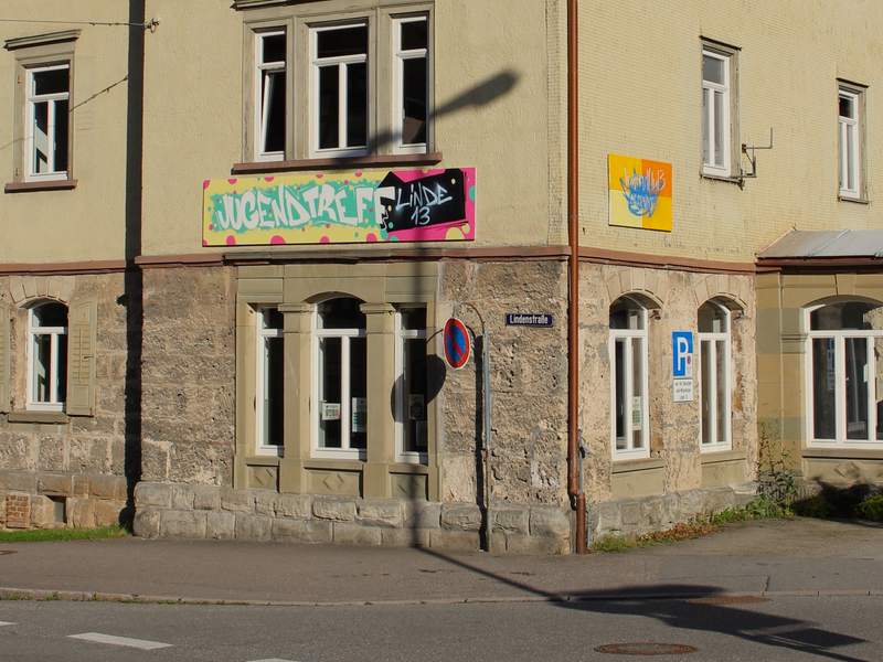 Zu sehen ist ein Teil eines Hauses. Auf der Hauswand ist ein Graffiti-Schild mit dem Schriftzug Jugendtreff Linde 13 zu sehen.