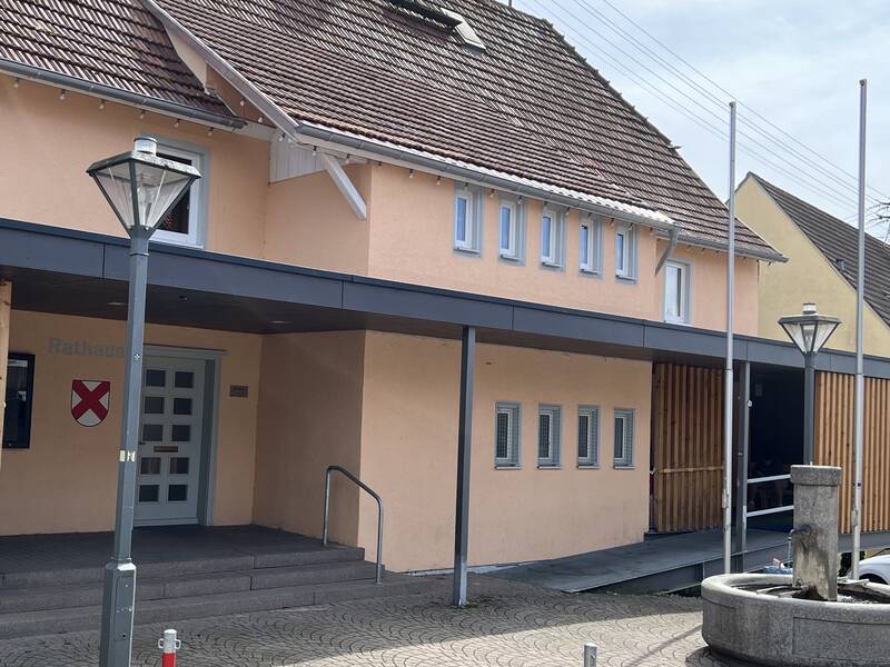 Zu sehen ist das Gebäude des Rathaus Bochingen, mit Bochinger Wappen am Eingang. Ein Brunnen und ein barrierefreier Zugang ist zu sehen.  