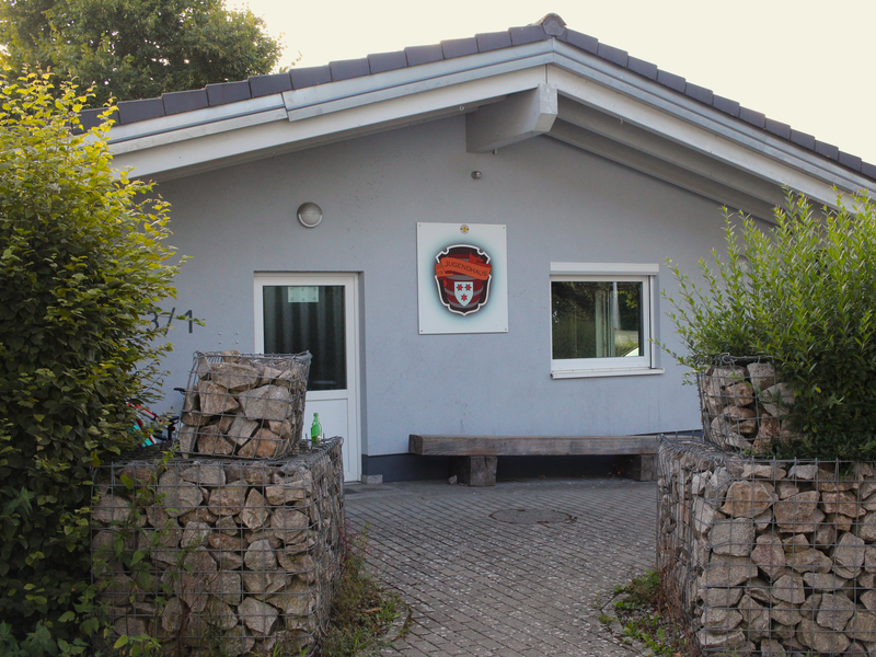 Vorderseite eines einstöckigen Gebäudes mit zwei Fenster und einer Türe. Das Jugendclubloge ist mit drei roten Sternen und einem Schriftzug mit Jugendclub ist zu sehen. Eine Holzbank steht vor dem Gebäude. Der Vorplatz ist gepflastert und mit zwei Steinmauern versehen. 