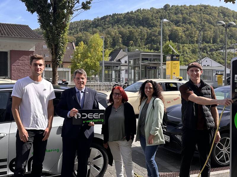 Drei Männer und zwei Fragen stehen vor einem eAuto mit Aufschrift deer und neben einer Aufladesäule. In der Hand. Bürgermeister Winter hält ein Schild mit Aufschrift deer. Im HIntergrund ist der Bahnhof von Oberndorf zu sehen.  